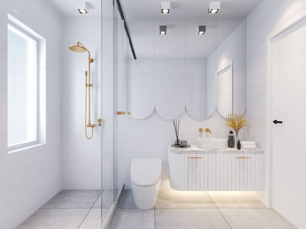 Bright contemporary bathroom with white tiled walls, large pale grey floor tiles, a frameless glass shower enclosure, wall-mounted rainfall shower in a gold finish, wall-hung toilet, and a fluted white vanity with a vessel basin. Arched scalloped mirror panels and warm under-vanity lighting add decorative detail.