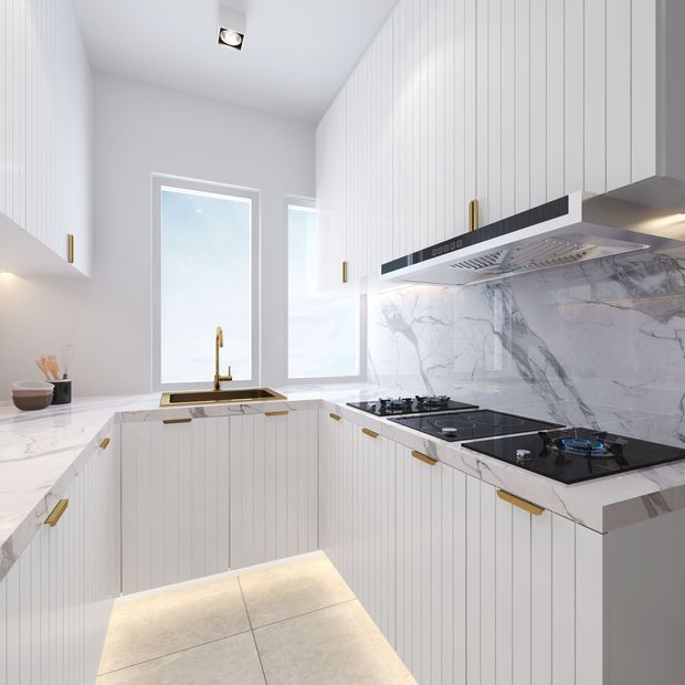 Bright contemporary galley-style kitchen with white vertically grooved cabinetry, slim brass handles, white marble-look benchtops and full-height marble-look splashback. A black gas cooktop and integrated rangehood occupy the right side, while a brass-toned sink tap sits beneath tall windows. Large pale floor tiles and under-cabinet lighting create a clean, luminous interior.