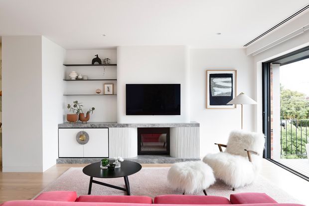 Bright contemporary living room with a white built-in media wall, open display shelves, wall-mounted television, and a linear fireplace set into fluted cabinetry. A black round coffee table, pale rug, pink sofa, and two white furry lounge chairs furnish the room. Large corner glazing opens to a balcony with greenery beyond.