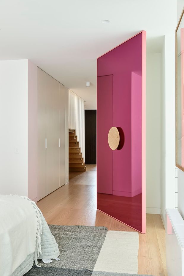Bright contemporary bedroom view toward a hallway, featuring a bold magenta pivot door with a round timber handle, pale timber flooring, white built-in wardrobes, a textured grey-and-cream rug, and a timber staircase with vertical slatted detailing in the background. The room is airy and playful with strong colour contrast and clean built-in architecture.