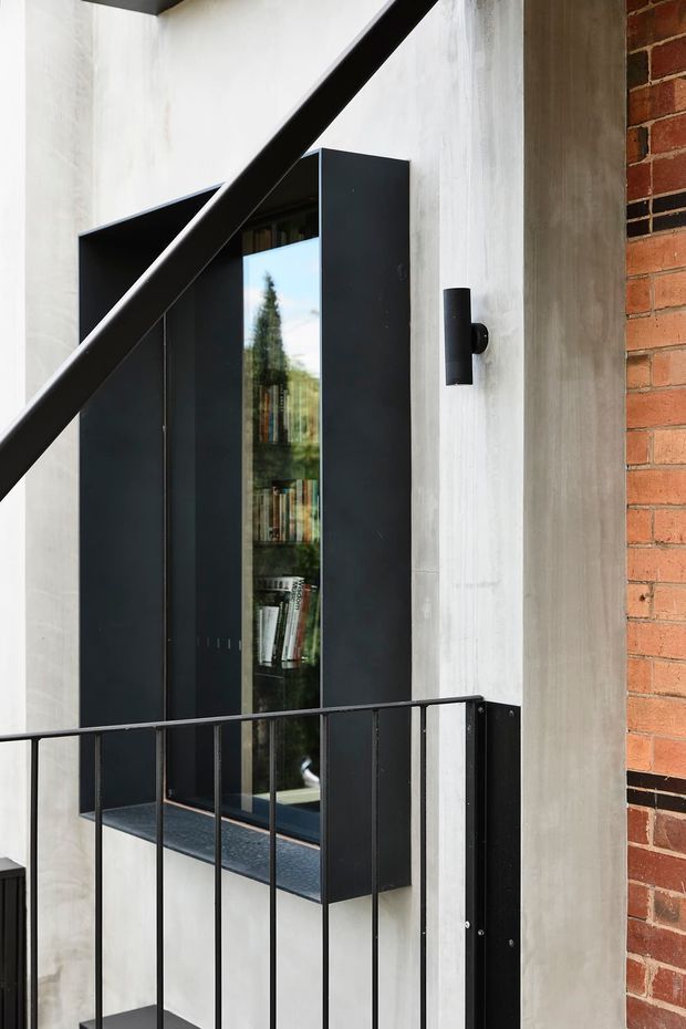 Close-up architectural view of a contemporary industrial-style facade or entry area. A tall matte-black built-in cabinet or window surround with reflective glazing and visible books sits against a white textured concrete/plaster wall. Black metal balustrading and a diagonal structural beam cross the foreground, while exposed red brick and a cylindrical black wall light add contrast.