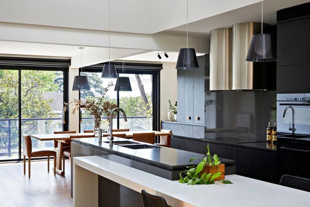 Contemporary open-plan kitchen and dining area with a large two-tier island, dark cabinetry, integrated appliances, reflective dark splashback, pendant lighting, and timber dining furniture. Full-height glazing brings in abundant daylight and overlooks leafy surroundings.
