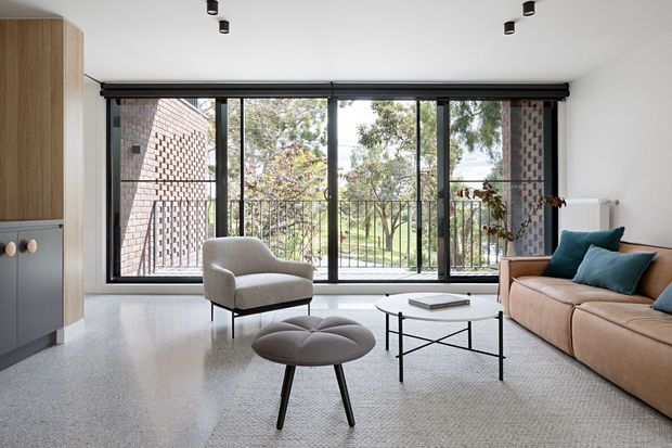 Bright contemporary living room with floor-to-ceiling black-framed glazing opening to a balcony, overlooking greenery. The room features a beige sectional sofa with teal cushions, a light upholstered armchair, a round coffee table, a sculptural ottoman, pale carpet, white walls and ceiling, and integrated light timber cabinetry with dark lower panels. Exposed brick is visible on the adjacent exterior wall.