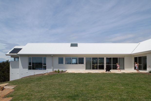 A wide exterior view of a contemporary single-storey home on a gently sloping landscaped site. The house has a long low profile, a large light-coloured pitched roof with skylights, white rendered or masonry walls, extensive dark-framed glazing, and a covered terrace opening onto a lawn. People and a dog provide scale.
