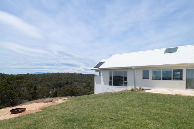 A single-storey contemporary house viewed from the rear or side, set on a gently sloping landscaped site overlooking dense native bushland. The white rendered exterior has a broad white corrugated pitched roof with a small skylight, extensive glazing, and a pale paved terrace. A lawn and simple fire-pit area occupy the foreground, with expansive sky and forest beyond.