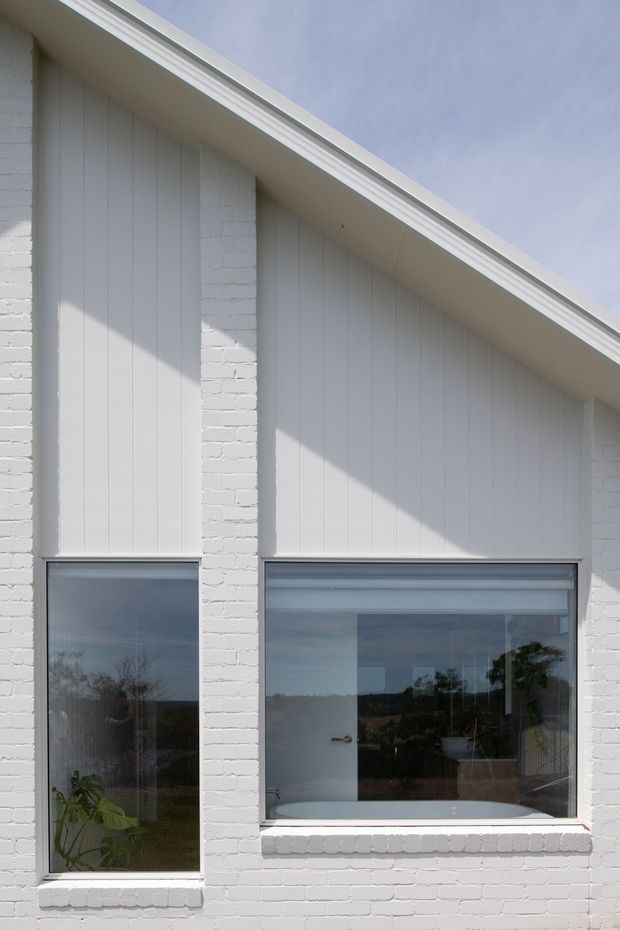 Close-up exterior view of a white-painted brick building with vertical white cladding beneath a sharply sloping gabled roof. Two large windows are visible; the right window reveals a freestanding bathtub inside, while the left shows indoor planting and reflections. The image emphasizes the clean white facade, window proportions, roof eaves, and mixed masonry/cladding exterior.