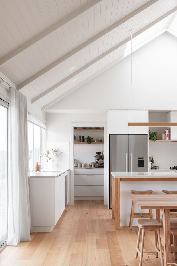 Bright, airy kitchen with a high pitched white timber-lined ceiling and exposed beams. The room features pale timber flooring, white cabinetry, a large stainless-steel refrigerator, open shelving, a white island with a timber-fronted breakfast bar, and timber stools. Full-height glazing with sheer curtains brings in abundant natural light.