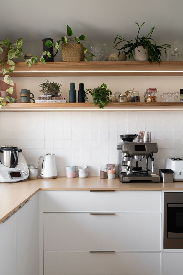 A bright, compact contemporary kitchen with pale cabinetry, light timber benchtops, and a white square-tile splashback. Open timber shelves with integrated strip lighting display potted plants, ceramics, glassware, books, and small kitchen accessories. The benchtop includes a premium espresso machine, kettle, multifunction cooker, toaster, and storage drawers, creating a warm, styled coffee and preparation area.