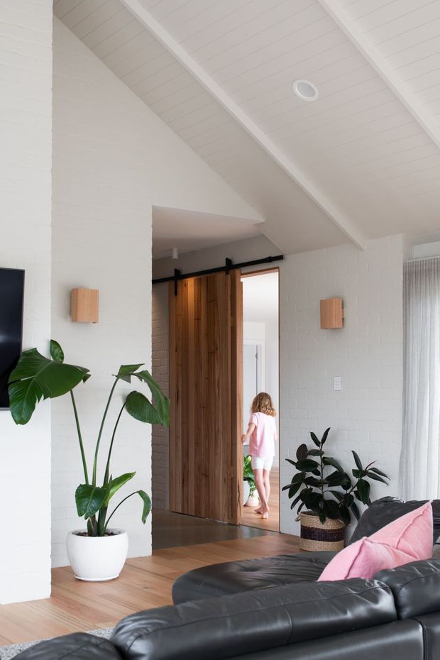 Bright contemporary living room with white-painted brick walls and a white-lined pitched ceiling. A large black leather sectional occupies the foreground, complemented by pink cushions, potted greenery, and simple timber wall sconces. A tall timber barn-style sliding door opens toward an adjoining space, with warm timber flooring throughout and sheer curtains at the window.