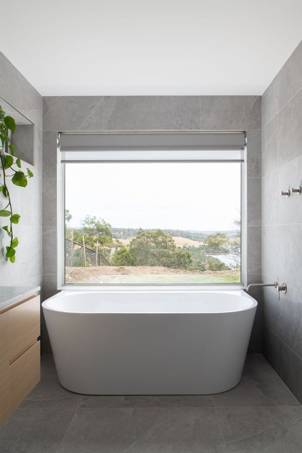 A contemporary bathroom centered on a freestanding white bathtub positioned beneath a large window with a roller blind and expansive landscape view. The room has full-height large-format grey stone-look wall and floor tiles, a light timber vanity with drawers at left, minimalist chrome tapware, and a small leafy plant. The composition is bright, calm, and clean with strong indoor-outdoor visual connection.