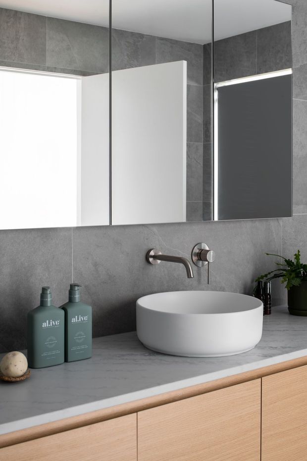 Contemporary bathroom vanity with a round white vessel basin, wall-mounted brushed metal tapware, grey stone-look tiled walls and benchtop, frameless mirrored cabinet, and light timber built-in cabinetry. Minimal greenery and bathroom accessories sit on the vanity.