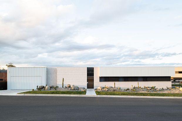 A wide exterior photograph of a low-profile contemporary building viewed across a paved street. The facade combines pale vertical metal cladding, light masonry or concrete block, large white horizontal wall panels, and a dark recessed strip of glazing. A minimalist gravel landscape with rocks and sparse cacti lines the frontage, beneath an overcast sky.