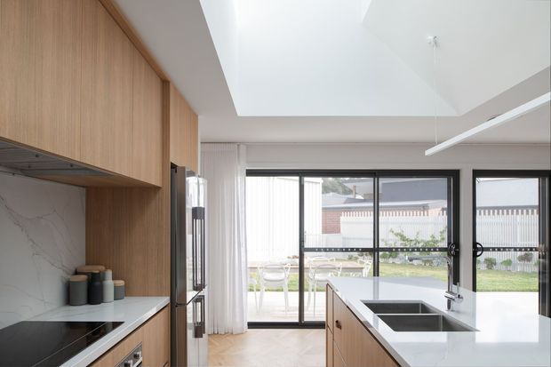 Bright contemporary kitchen with extensive vertical-grain timber cabinetry, a white veined stone splashback and benchtops, integrated appliances, and a central island with a double dark sink. Full-height black-framed glazing opens toward an outdoor dining and garden area, while a large skylight or raised ceiling zone increases natural light. The space has a clean, refined, warm-modern appearance.