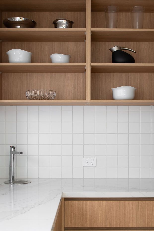 Close-up view of a refined kitchen fit-out featuring open, vertical-grain timber shelving with ceramics and glassware, a white square-tile splashback, pale veined stone benchtop, integrated timber cabinetry, and a slim chrome tap. The composition is clean and understated with warm timber contrasting against white surfaces.