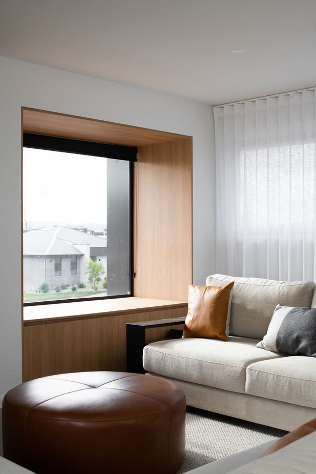 A bright, compact living room with a large built-in timber-lined window seat and deep timber reveal framing a black-framed window. A light beige upholstered sofa with grey and tan cushions faces a round brown leather ottoman, with a small black side table and neutral rug. Sheer white curtains filter daylight, creating a calm contemporary interior.