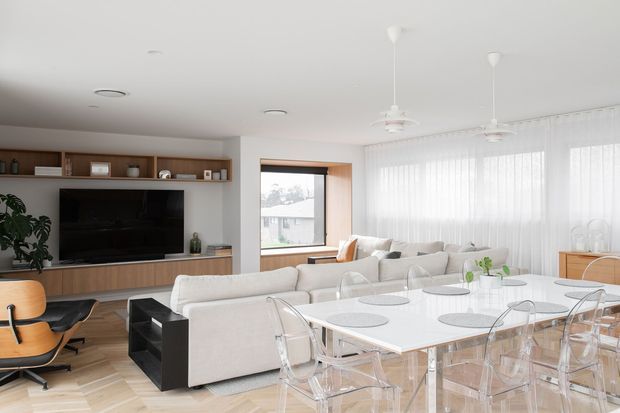 Bright open-plan living and dining room with a contemporary, mid-century-influenced fit-out. The space features white upholstered sofas, a large white dining table with transparent acrylic chairs, warm timber herringbone flooring, a built-in timber media wall, extensive sheer-curtained glazing, and sculptural pendant ceiling fans.