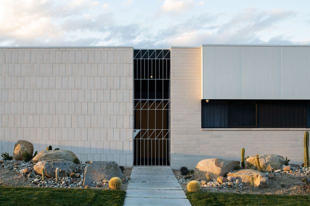 Single-storey contemporary building facade with a geometric composition of pale masonry and smooth light-grey cladding, a tall narrow glazed entrance, and a recessed dark horizontal window band. A concrete path leads through a xeriscaped front garden with boulders, gravel, and sculptural desert planting.