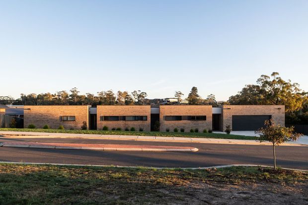 Street-facing view of a low, single-storey contemporary residential building composed of several rectilinear brick-clad volumes. Long horizontal dark-framed windows and recessed openings punctuate the warm brick facade, with a dark garage door at the right. The building sits behind a paved footpath and landscaped verge, with mature trees in the background and warm late-afternoon light.