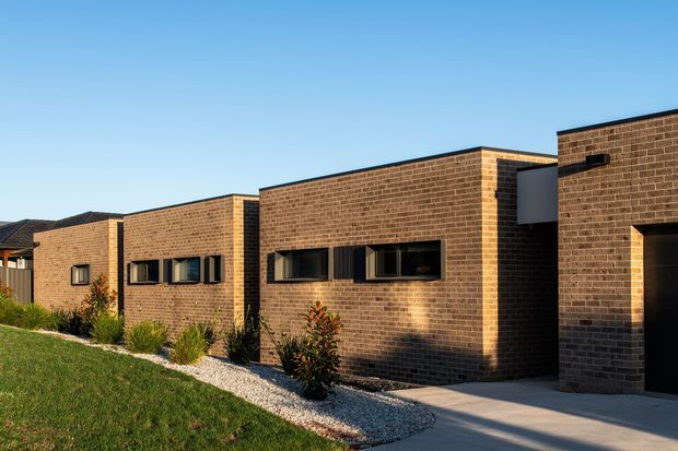 Exterior view of a contemporary single-storey residential building composed of several stepped rectangular brick volumes. The facade features warm brown brickwork, dark-framed horizontal windows, a flat roofline, and a concrete driveway or path, with landscaped planting and lawn in the foreground.