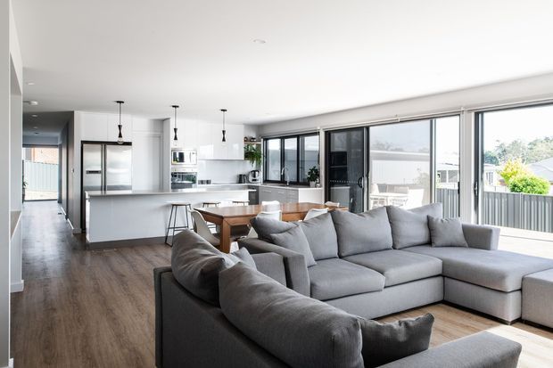 Bright open-plan living area with a large grey sectional sofa in the foreground, dining table behind it, and a contemporary white kitchen with island, integrated appliances, and pendant lights. Full-height black-framed glazing opens the room to an outdoor balcony or terrace, while dark timber-look flooring runs throughout.