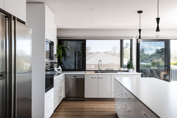 Bright contemporary kitchen with white full-height cabinetry, stainless-steel appliances, a large central island, timber-look flooring, expansive black-framed windows, and minimalist pendant lights. The kitchen overlooks a leafy suburban setting.
