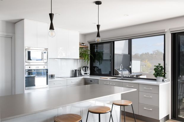 Bright contemporary kitchen with glossy white full-height cabinetry, integrated microwave and oven, a large grey island with timber-topped stools, dark lower cabinetry, and expansive black-framed windows overlooking trees. Minimal pendant lights and indoor plants add restrained warmth.