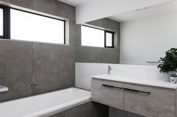 Contemporary bathroom featuring a white built-in bathtub, large grey concrete-look wall tiles, a floating grey vanity with black linear handles, white benchtop and integrated basin, black-framed horizontal windows, and a large wall mirror. The space has a clean, minimalist fit-out with neutral tones and natural light.