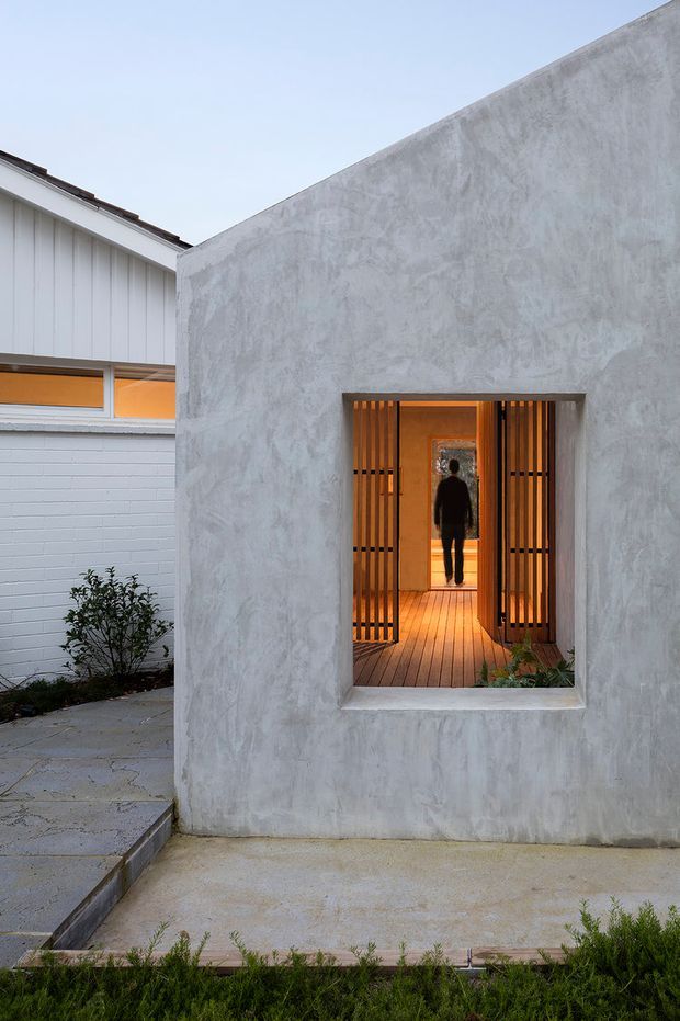 Exterior view of a compact contemporary dwelling or addition, focused on a tall pale grey textured rendered wall with a deeply recessed entry opening. Warm timber decking and vertical timber slatted gates frame the doorway, while an older white weatherboard and brick structure is visible to the left. Concrete paving, a small planted courtyard edge, and warm interior lighting are visible through the entrance.