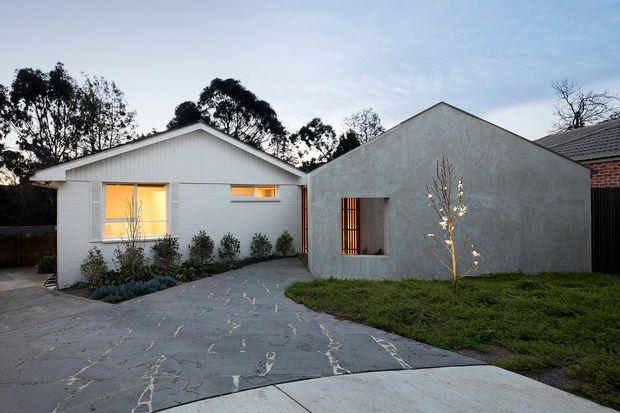 Single-storey residential building with two intersecting gabled volumes. The left volume has white vertical corrugated cladding, a large illuminated window, a narrow horizontal window, and a garage-style door, while the right volume has a textured grey rendered facade and a deeply recessed timber-framed opening. A cracked concrete driveway and sparse landscaped planting occupy the foreground, with neighbouring residential fabric visible at the edges.