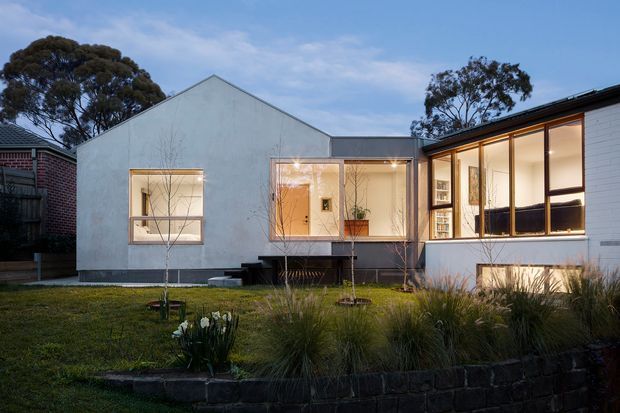 Front exterior view of a contemporary single-storey residence at dusk. The home has a simple gabled central volume joined to a lower glazed wing, with pale rendered walls, dark metal roof edging, broad timber-framed windows and a raised entry connection. A landscaped front garden with grasses and a low dark masonry retaining wall sits in the foreground.
