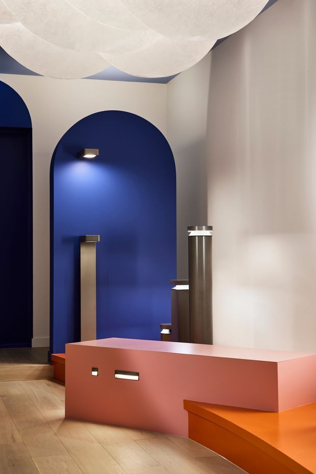 A stylised interior feature space with a deep cobalt-blue arched alcove, sculptural metallic cylindrical elements, integrated lighting, and a large coral-pink/orange built-in counter or display structure. The room has pale walls, warm timber flooring, and an oversized layered white sculptural ceiling light. The composition is bold, geometric, and highly curated, resembling a contemporary commercial or hospitality interior.