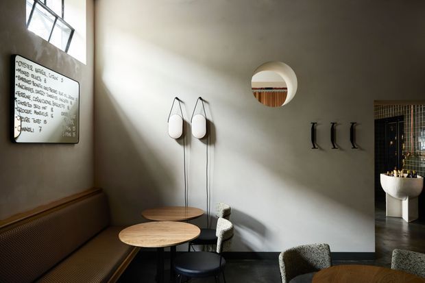 A moody, warmly lit contemporary café or hospitality interior with a long built-in upholstered bench along a textured plaster wall, small round timber tables, dark chairs, sculptural pendant lights, and black coat hooks. A circular wall opening reflects or reveals timber detailing, while an open doorway leads to a tiled washroom area with a wall-mounted basin. A handwritten menu board is mounted near the high window.