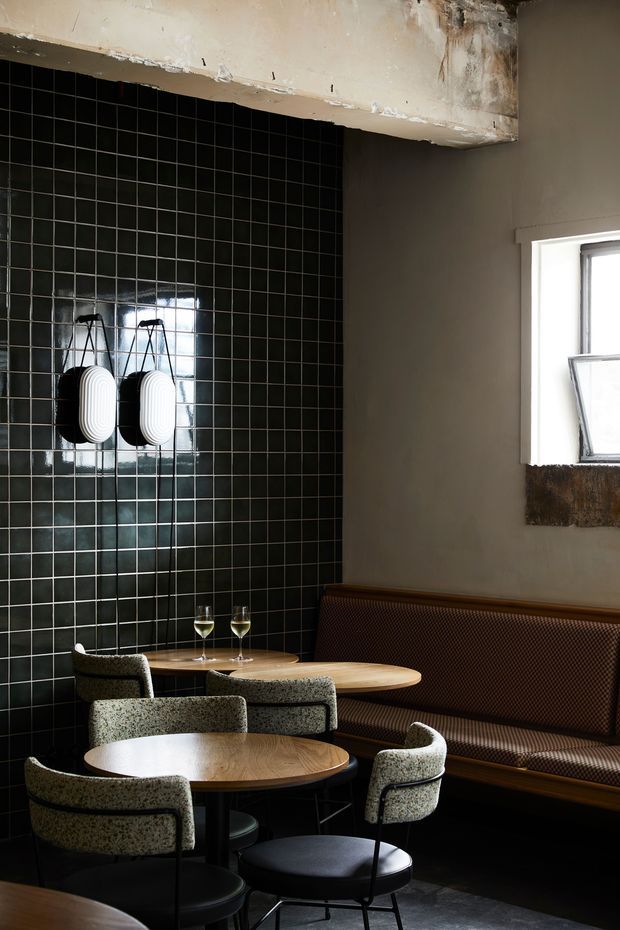 A moody hospitality dining area with a long upholstered banquette, several round timber tables, and sculptural upholstered chairs. The space features a dramatic glossy black tiled feature wall, exposed rough concrete and plaster overhead, dark flooring, and a small industrial-style window with a deep timber sill. Two pendant lights hang against the tiled wall, while wine glasses and table settings suggest a restaurant or café interior.