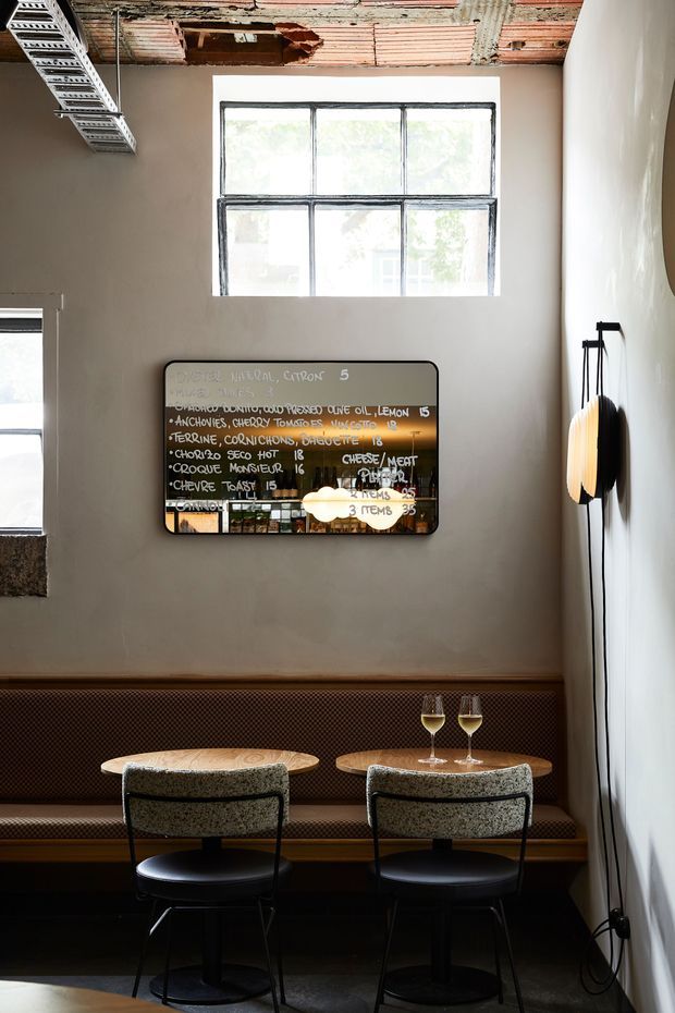A compact contemporary hospitality-style dining nook with two small round tables, upholstered bar-height chairs, and a long built-in banquette along the wall. A mirrored menu board hangs above the seating, while tall industrial-framed windows bring in daylight. The space combines dark flooring, warm timber accents, textured upholstery, and an exposed unfinished ceiling with visible services and ductwork.