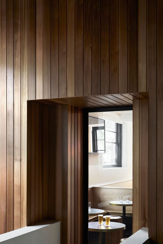 A close architectural interior view framed by richly stained vertical timber wall panelling. A timber-lined opening reveals a bright adjoining room with black-framed windows, pale walls, booth-style seating, and small round tables. The composition emphasizes crafted joinery, contrasting dark wood, and a warm, intimate atmosphere.
