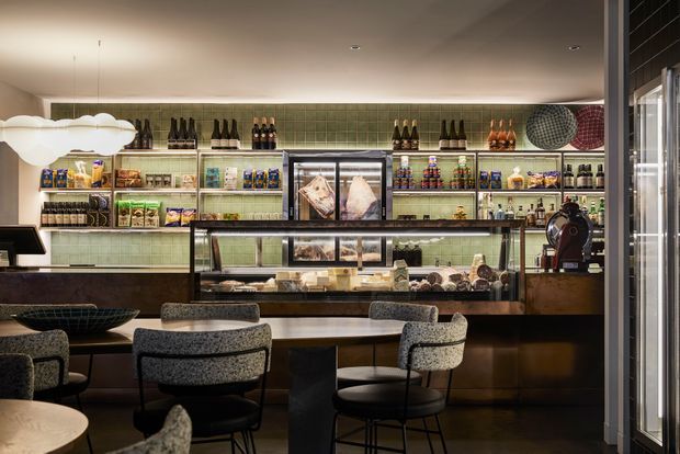 A refined contemporary café or deli interior viewed from the dining area toward a long service counter. The space features a dark timber or stained-wood counter, glass pastry display cases, open shelving stocked with packaged goods and bottles, green tiled wall cladding, upholstered dining chairs, and sculptural pendant lighting. The palette is warm and subdued, with polished commercial detailing and a compact, intimate atmosphere.