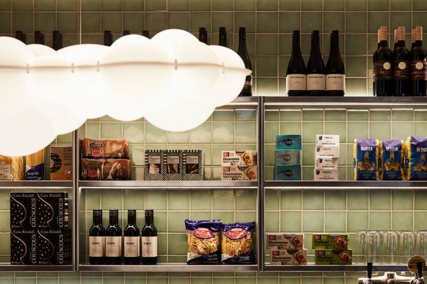 A compact, warmly lit retail or hospitality shelving display with dark green square tiled walls and open metal shelving. The shelves hold wine bottles, packaged food products, boxed goods, and rows of drinking glasses. A large glowing white cloud-shaped pendant light dominates the upper left, creating a playful focal point against the orderly commercial-style display.