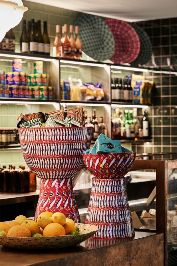 A richly styled commercial bar or food-service interior with dark green tiled walls, illuminated glass-front shelving stocked with bottles, canned goods and pantry products, and a polished dark counter in the foreground. Two prominent sculptural display bowls or pedestal vessels feature bold red, white and blue patterned surfaces, with a fruit bowl of oranges and limes beside them. Large circular patterned decorative panels hang above the shelving, creating a colourful, eclectic atmosphere.