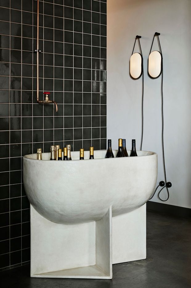 A dramatic, sculptural bathroom interior featuring a large freestanding white vessel bathtub on a pedestal base. The left wall is clad in glossy black square tiles with copper plumbing and a wall-mounted shower fixture. Two oval pendant wall lights with black cords hang against a pale plaster wall, while dark flooring and bottles placed in the tub add contrast.
