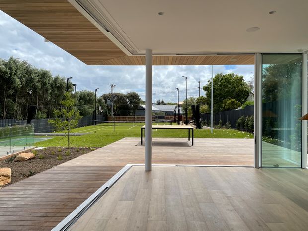A contemporary indoor-outdoor living space opens through large sliding glass doors onto a broad timber deck and landscaped backyard. The covered terrace has a timber-lined soffit, slim white structural column, recessed ceiling lights, and pale wood-look flooring. Beyond the deck is a green lawn, planted garden beds, fencing, and a recreational court or play area.