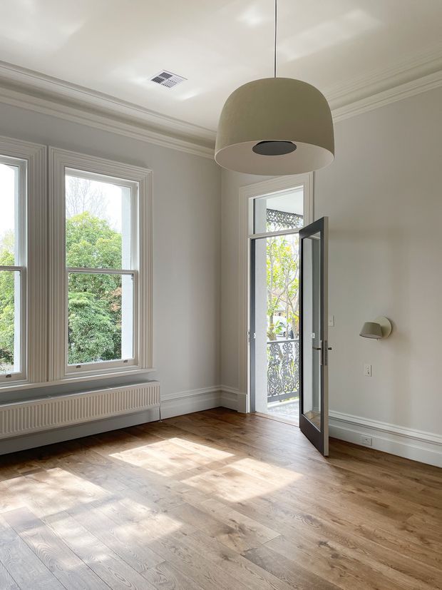 Bright, mostly empty traditional-style room with tall sash windows, ornate layered architraves and ceiling cornices, white-painted walls, and warm timber flooring. A large neutral pendant light hangs centrally, complemented by a small wall sconce. An open glazed door leads to a balcony or exterior landing, while sunlight creates broad patterns across the floor.