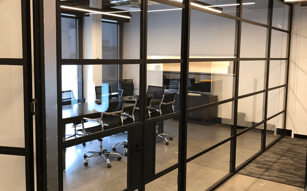 A modern office meeting room viewed through a black metal-framed glass partition. The room contains a conference table with rolling office chairs, computers, and a wall-mounted display, with polished concrete flooring, recessed linear lighting, and a neutral palette. A glazed corridor or adjacent workspace is visible to the right.