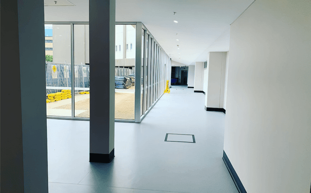 A bright, minimalist commercial or institutional corridor with a smooth pale floor, white walls, dark skirting, and dark structural columns. Full-height glazed partitions line one side, revealing an active construction or industrial area outside. Recessed ceiling lights and clean linear detailing create a spacious, contemporary interior.