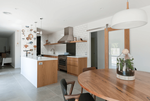 Bright open-plan kitchen and dining area with a timber island, white tiled splashback, stainless-steel rangehood and freestanding cooker, plus a round timber dining table in the foreground. White cabinetry and walls are balanced by warm timber joinery, exposed timber door framing, and contemporary pendant lighting.