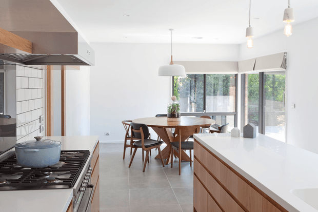 Bright open-plan kitchen and dining area with a large white island featuring timber drawer fronts in the foreground. The kitchen includes a gas cooktop, pale blue cookware, white tiled splashback, timber-toned cabinetry, and a contemporary rangehood. Beyond, a round timber dining table with mid-century-style chairs sits beside extensive glazed doors and windows, creating an airy connection to the outdoors. Pendant lights and neutral finishes give the space a clean, warm contemporary character.