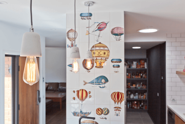 Bright contemporary kitchen interior with a decorative tiled feature wall depicting colourful hot-air balloons, airships and whimsical flying motifs. Pendant lights with exposed filament bulbs hang in the foreground, while a timber door, dark open pantry shelving, white subway-tile backsplash and light benchtop are visible around the space. A living area is partially visible beyond the doorway.