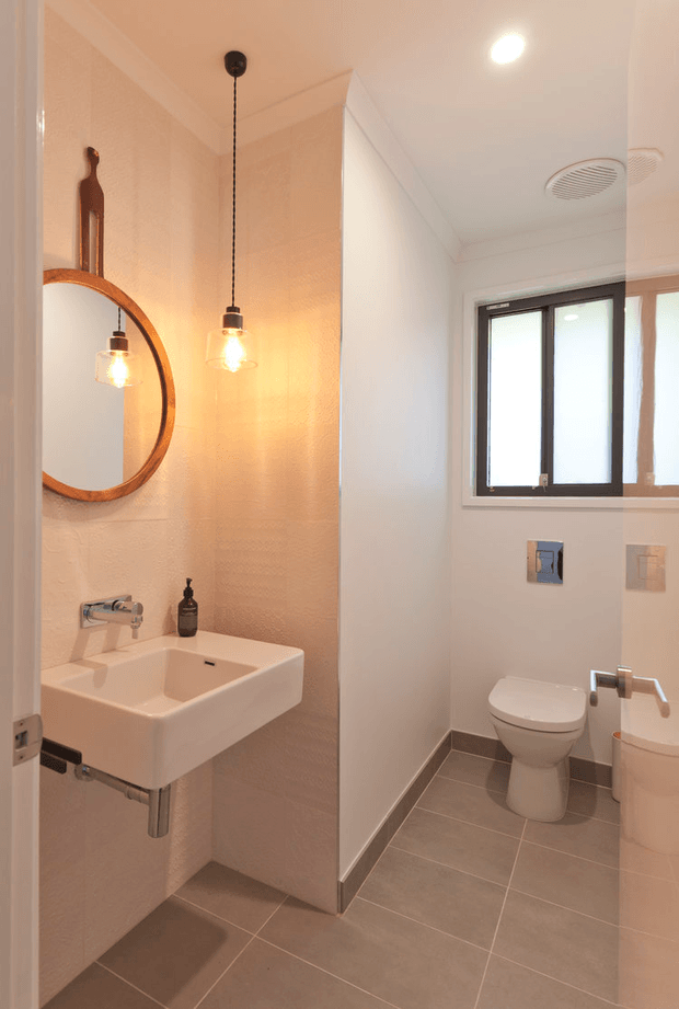 A compact contemporary bathroom with a wall-mounted white basin, exposed chrome plumbing, wall-mounted tap, round timber-framed mirror, and two warm pendant lights. A separate toilet area is visible beyond the dividing wall, with a wall-hung flush plate, frosted window, white toilet, and pale tiled floor. The room has white textured walls, grey-brown floor tiles, and warm neutral lighting.