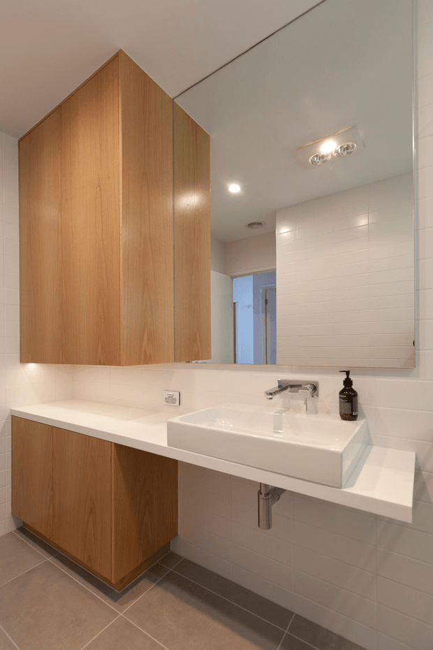 A compact contemporary bathroom vanity with warm timber cabinetry, a white integrated countertop and rectangular basin, chrome wall-mounted tapware, and a large mirrored cabinet. White tiled walls and a grey-beige tiled floor create a clean, practical interior with warm recessed lighting.