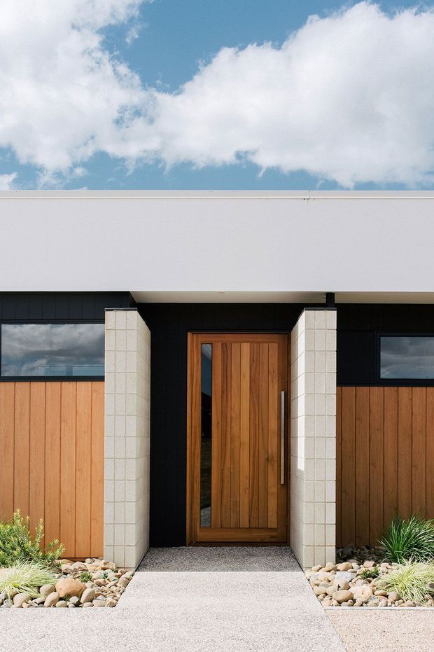 Front-facing view of a contemporary single-storey home entrance. A tall timber pivot door with a narrow glazed sidelight is framed by pale masonry block piers beneath a broad white flat roof overhang. Dark cladding and horizontal windows contrast with warm vertical timber wall panels. A textured concrete path leads through a low-maintenance rock garden with ornamental grasses.