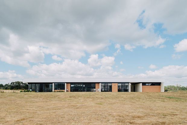 A long, low-slung single-storey contemporary building set in an open rural landscape. The facade combines extensive dark-framed glazing, pale rendered or concrete elements, warm timber-toned panels, and a broad shallow roofline. A dry grass field and expansive cloudy blue sky surround the building.